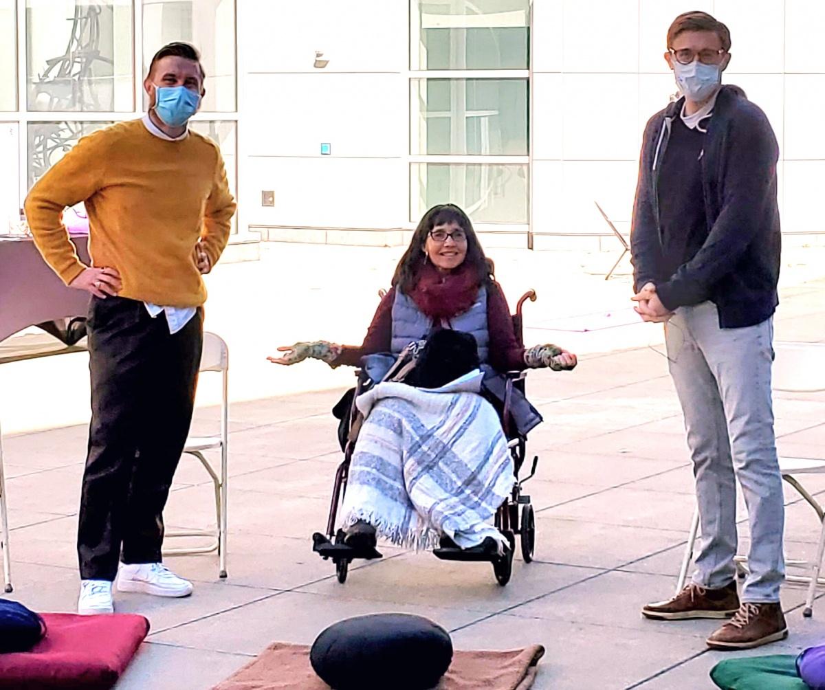 A women with shoulder-length brown hair sits in a wheelchair in the Crocker's outdoor courtyard, her hands outstretched and palms facing up. Tw men stand on either side of her in face masks, smiling. Cushions and blankets are on the ground in front of them.